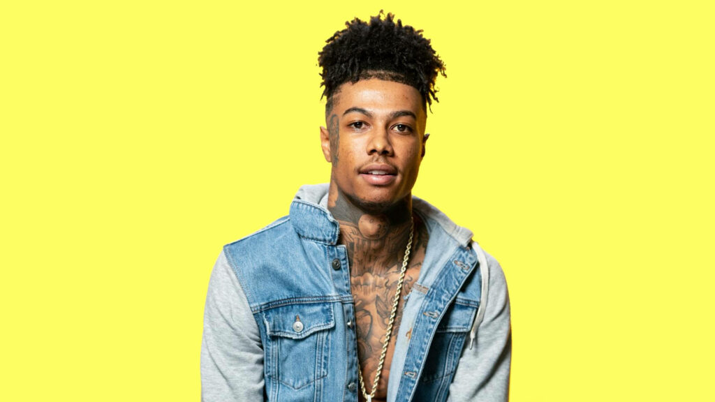 Blueface in denim vest and gray hoodie posing confidently on vibrant yellow wallpaper background