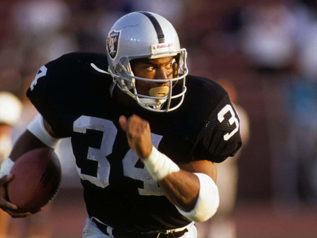 Bo Jackson running with football in Raiders uniform showing fierce determination in intense game Wallpaper