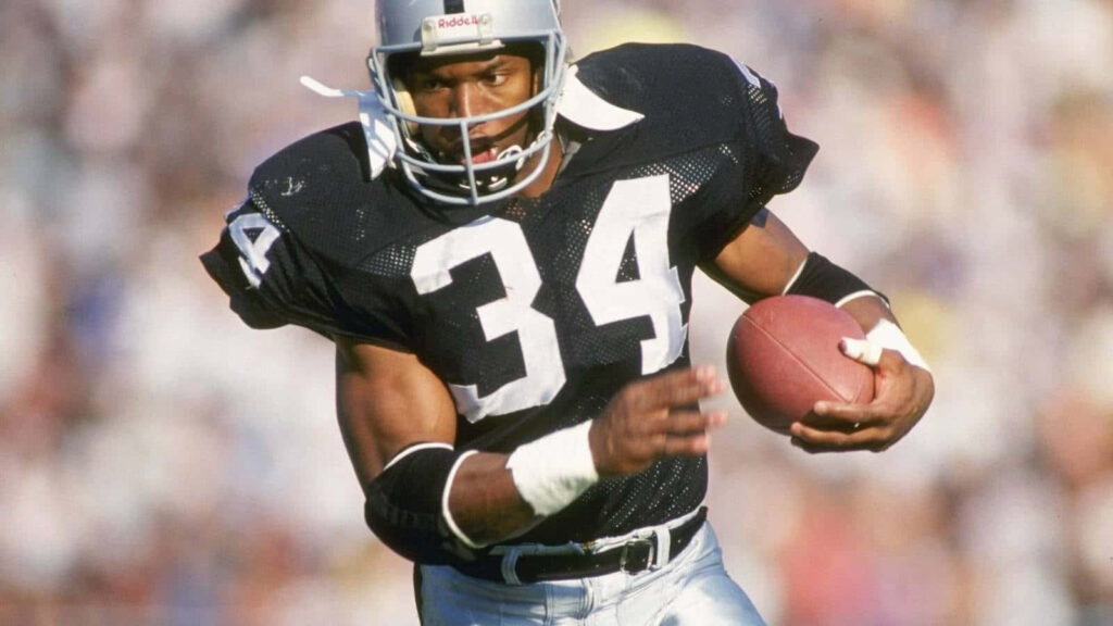 Bo Jackson charging forward in Raiders uniform gripping football action NFL wallpaper background