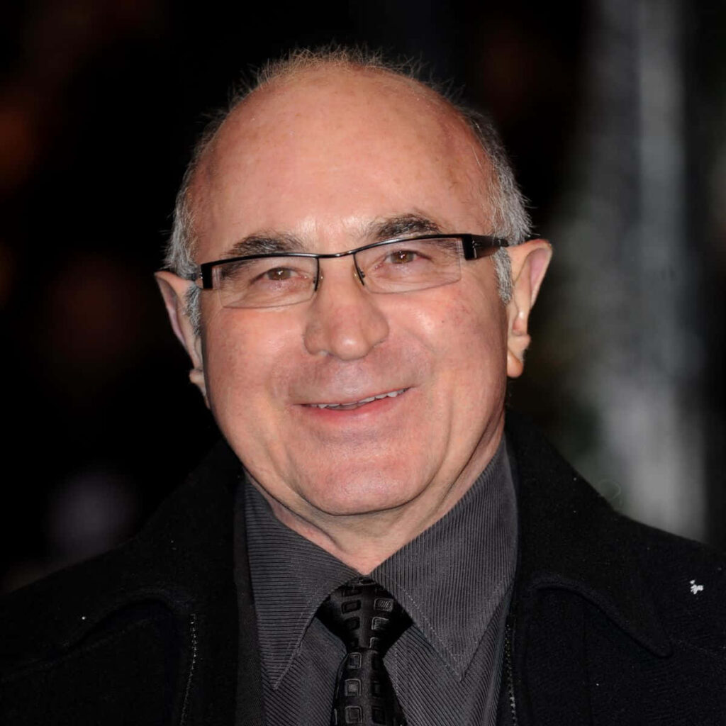Bob Hoskins in formal suit and glasses smiling warmly with blurred background wallpaper