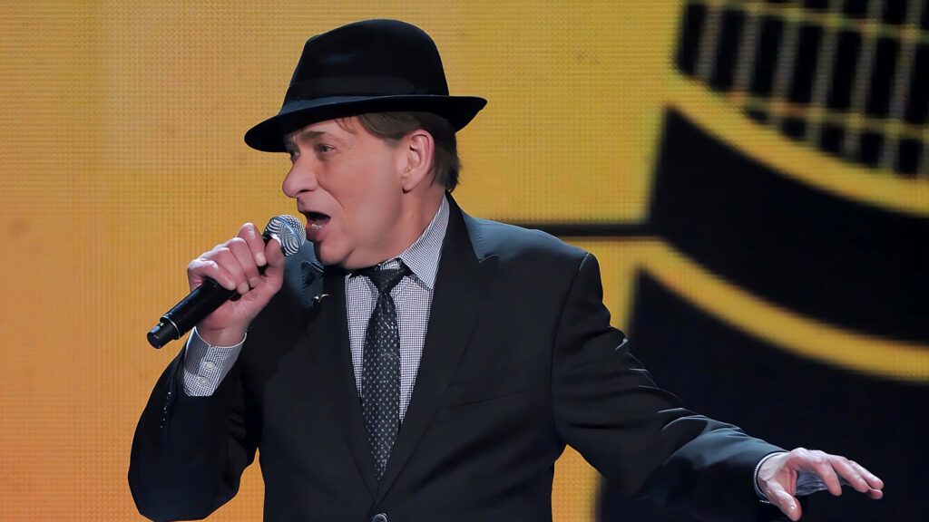 Bobby Caldwell live performing in classic suit and hat with vibrant wallpaper stage background