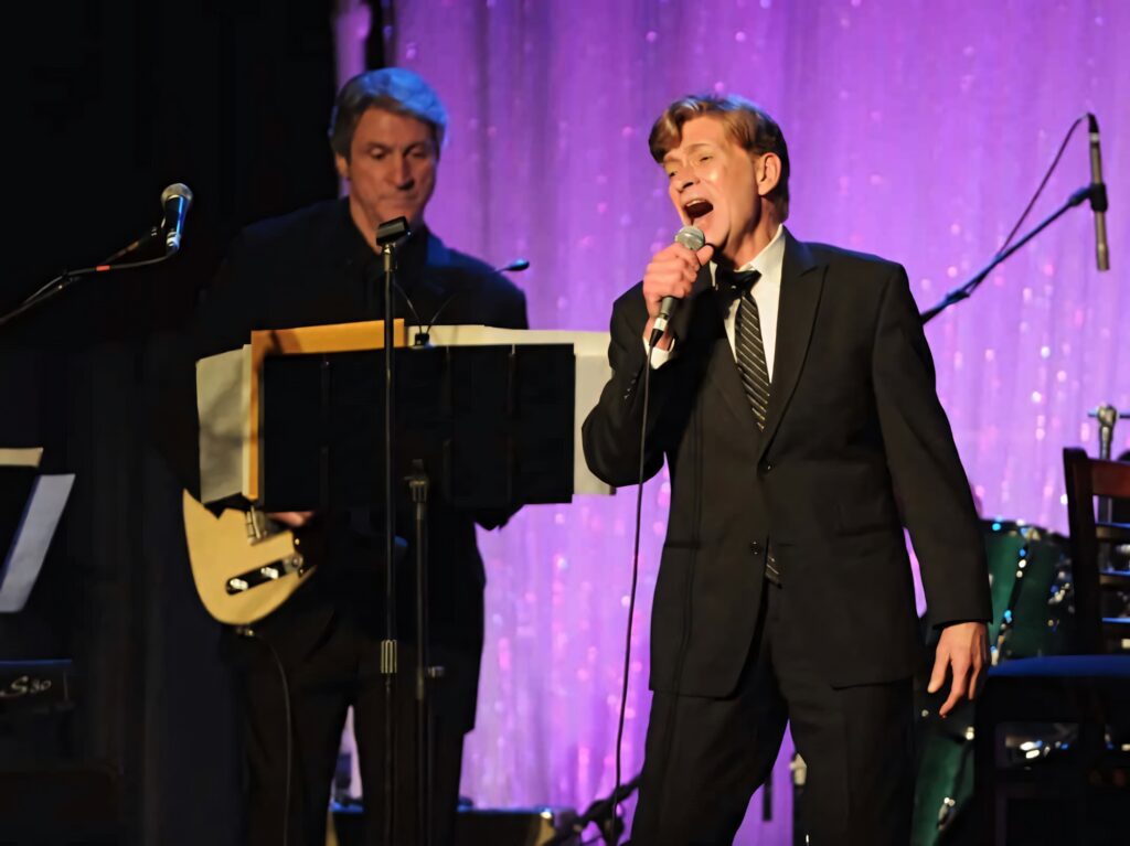 Bobby Caldwell singing live on stage in sharp suit with band and sparkling purple backdrop wallpaper