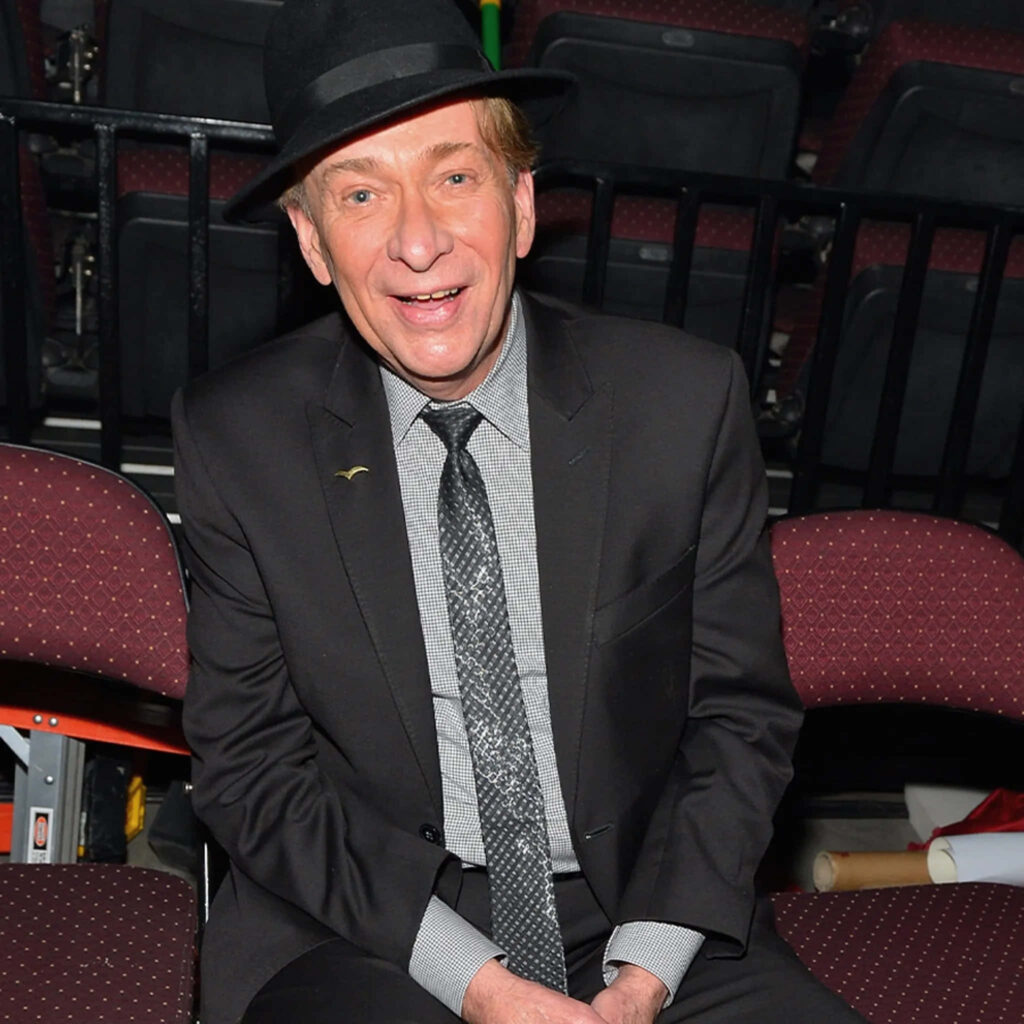 Bobby Caldwell in suit and hat performing live at concert venue with energetic wallpaper background
