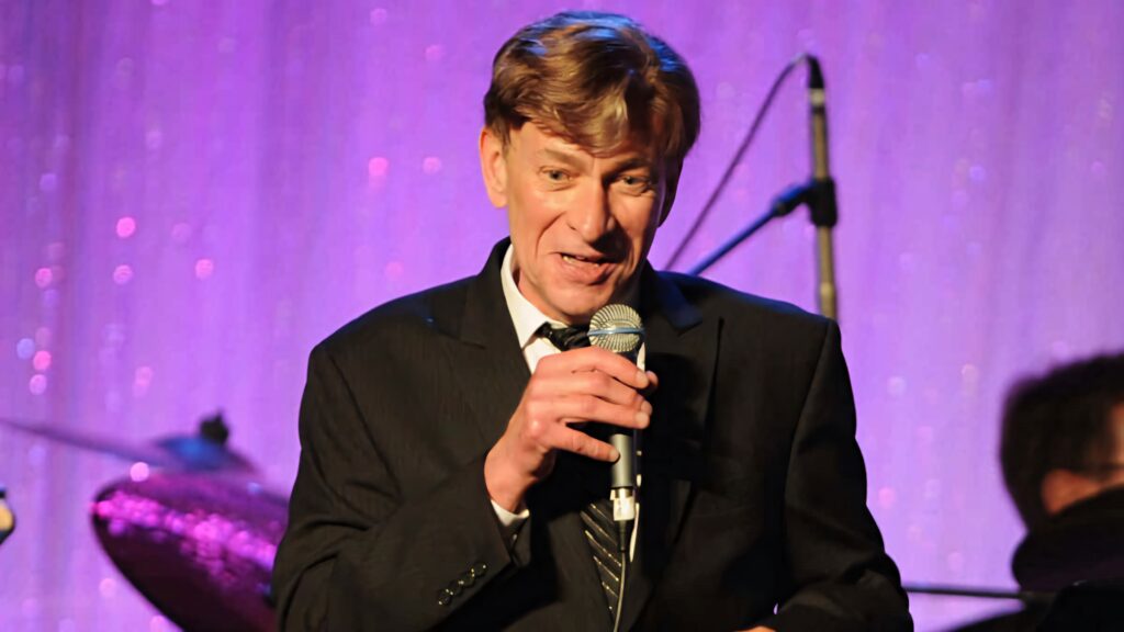 Bobby Caldwell live in suit singing with microphone vibrant stage lights and instruments background Wallpaper