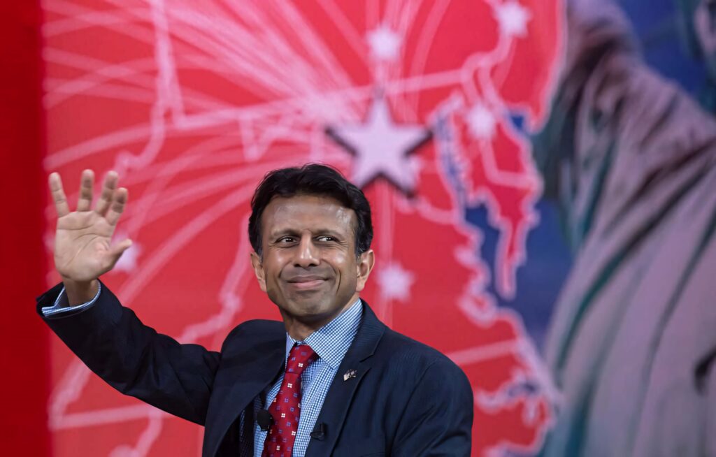 Bobby Jindal waving on stage with confident smile red patriotic backdrop wallpaper background