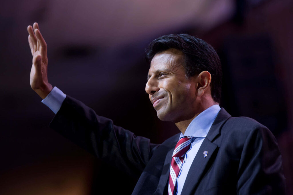 Bobby Jindal smiling waving on stage in formal suit and tie with wallpaper background