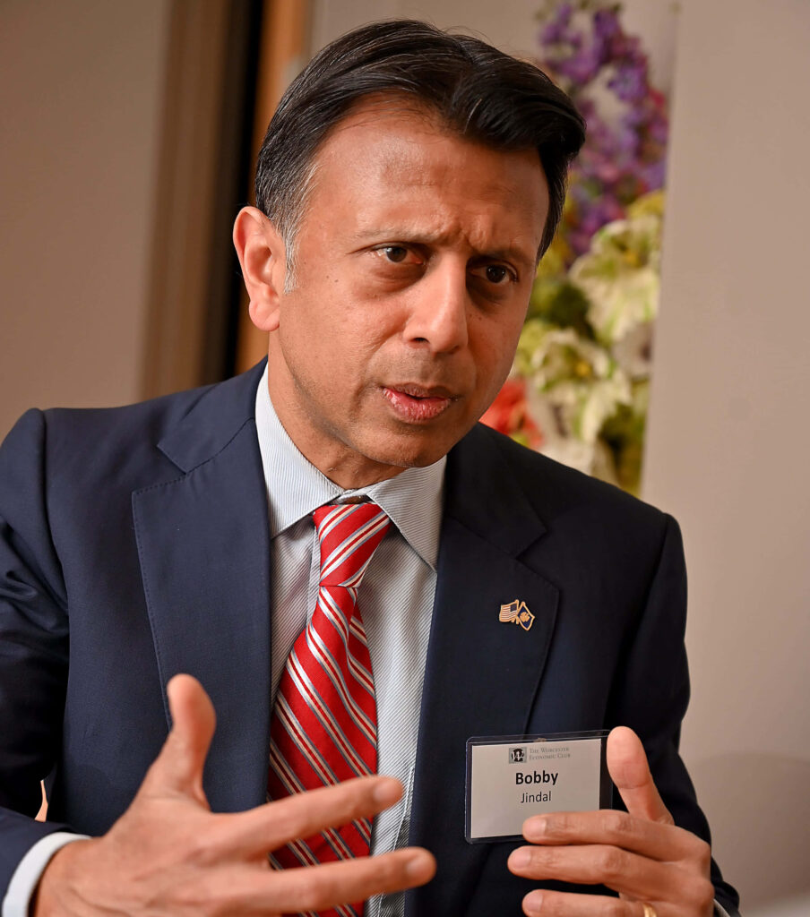 Bobby Jindal in suit with name badge speaking animatedly against wallpaper background