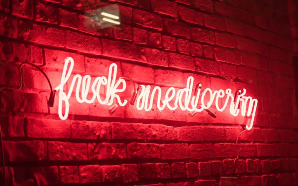 Bold cursive neon sign saying fuck mediocrity glowing on dark red brick wall wallpaper