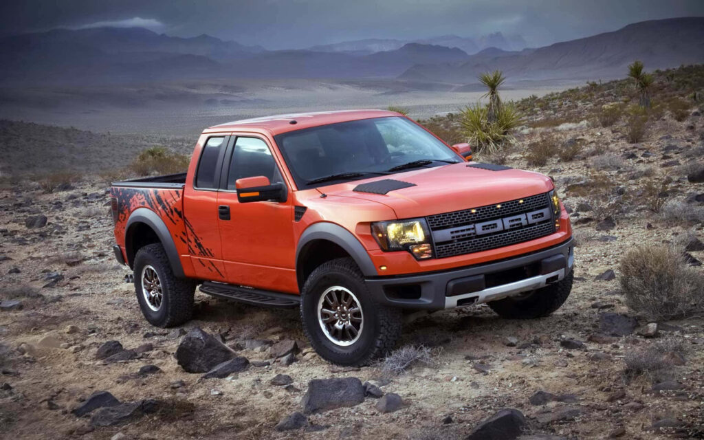 Orange Ford F-150 Raptor parked on rocky desert terrain under cloudy sky showcasing rugged off-road design Wallpaper