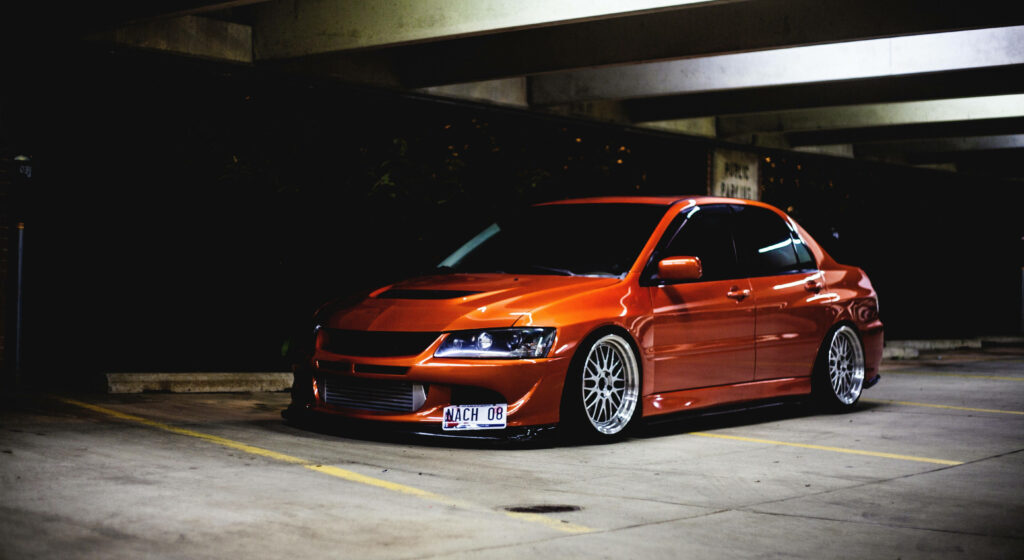 Striking orange JDM car with custom rims parked in dimly lit lot showcasing aggressive stance Wallpaper