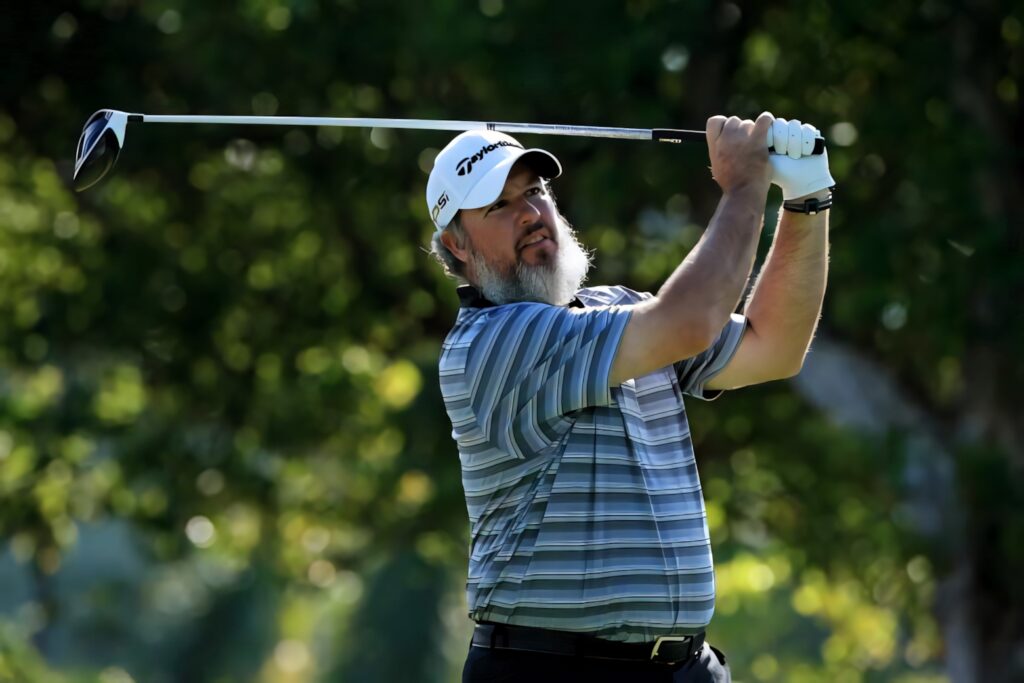 Grimacing Boo Weekley mid-swing on vibrant golf course with lush trees and sunlight background Wallpaper
