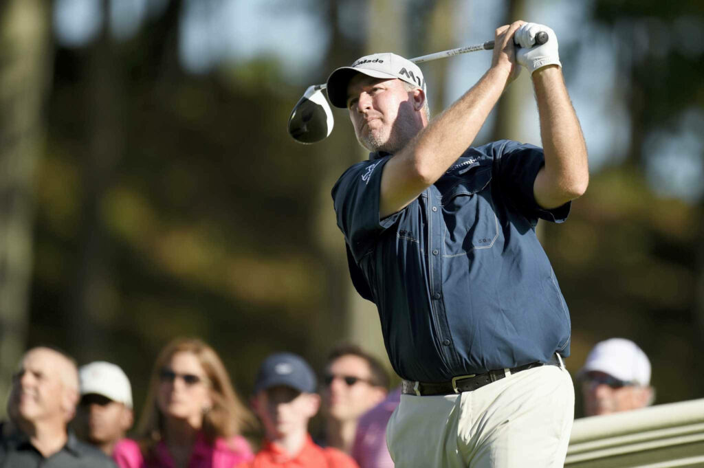 Boo Weekley golfing mid-swing in navy shirt with blurred audience on sunny golf course background Wallpaper