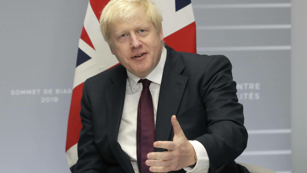 Boris Johnson in dark suit red tie seated before United Kingdom flag patriotic wallpaper background