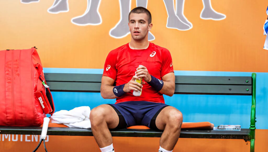 Borna Coric resting on bench during tennis break in red shirt blue shorts vibrant sports wallpaper