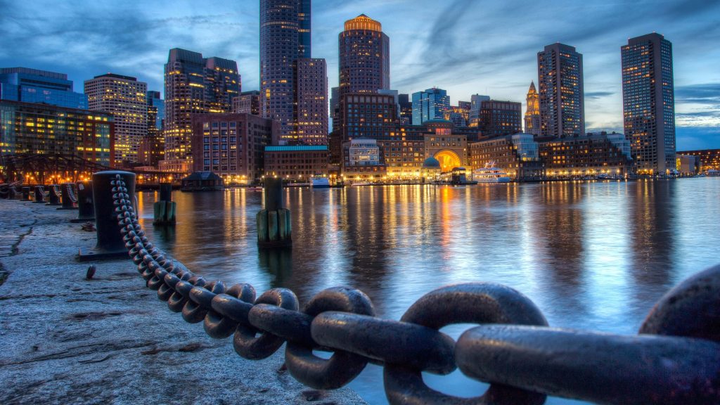 Rusty iron chain frames Boston skyline illuminated at twilight with golden lights on calm harbor water Wallpaper