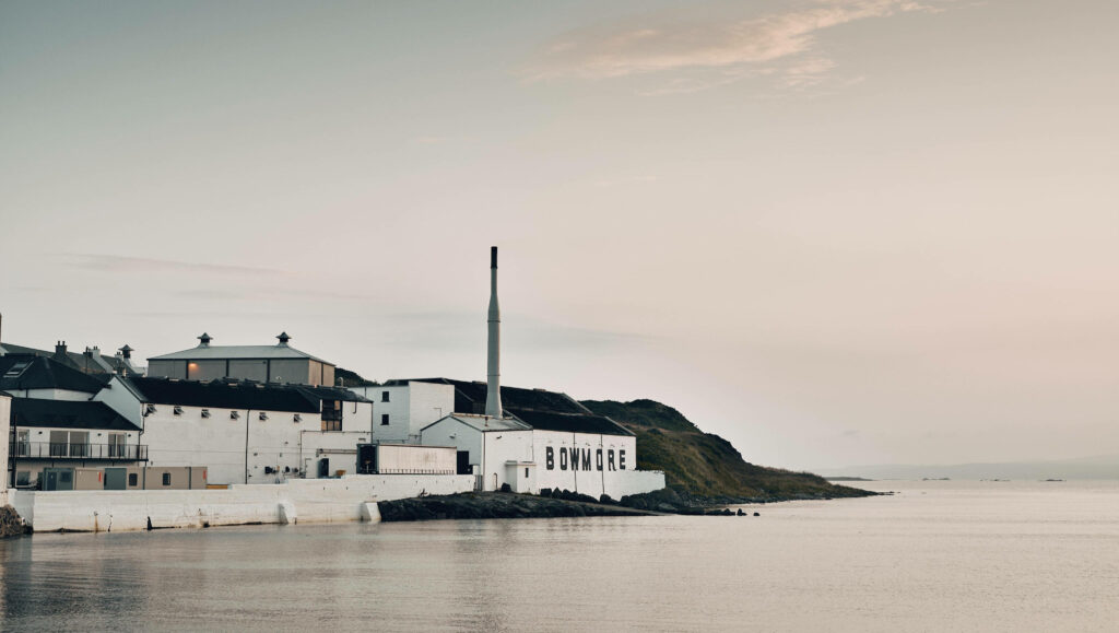 Bowmore Islay distillery whitewashed buildings and smokestack by calm coastal waters at sunset Wallpaper