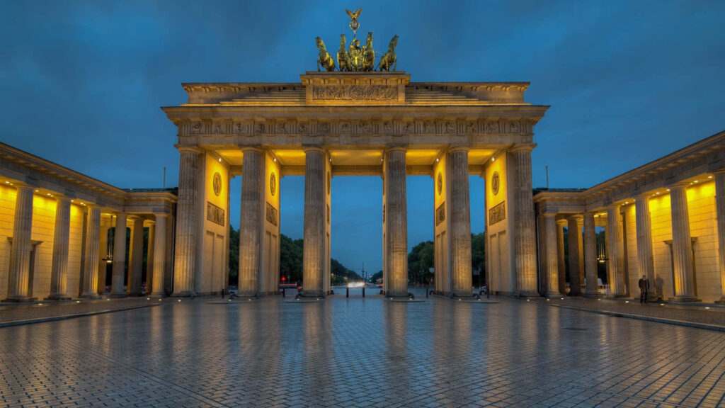 Brandenburg Gate Berlin illuminated at dusk with golden lights and reflective cobblestone plaza Wallpaper