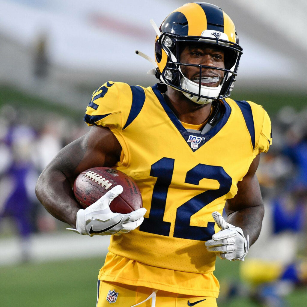 Brandin Cooks jogging with football in vibrant Los Angeles Rams uniform on dynamic field background Wallpaper