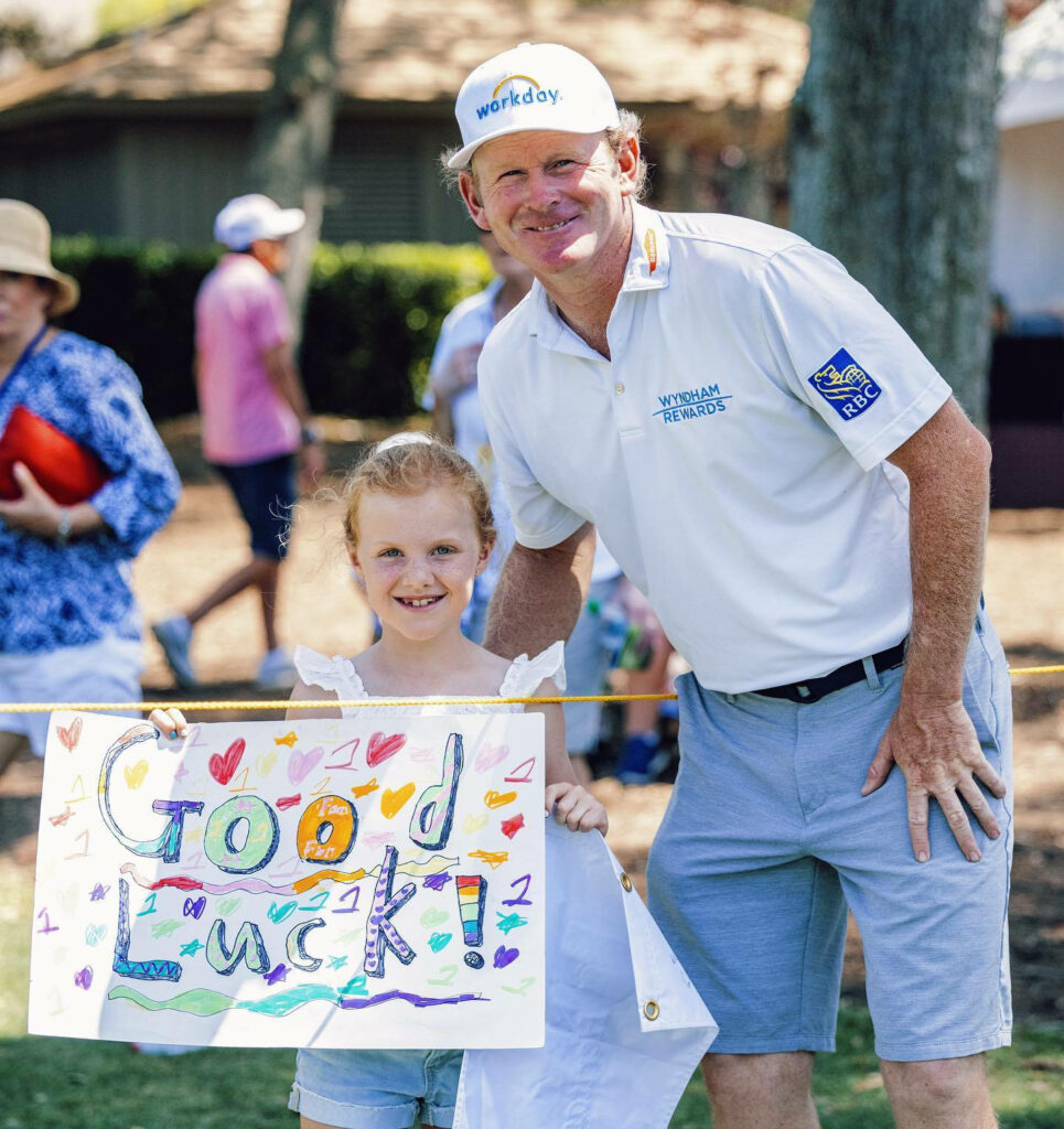 Brandt Snedeker smiling with young fan holding colorful good luck sign at sunny golf event Wallpaper
