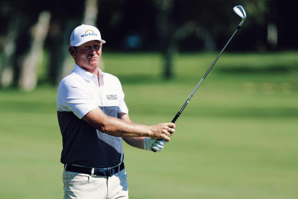 Brandt Snedeker mid-swing on sunlit golf course wearing white cap and striped polo shirt golf wallpaper