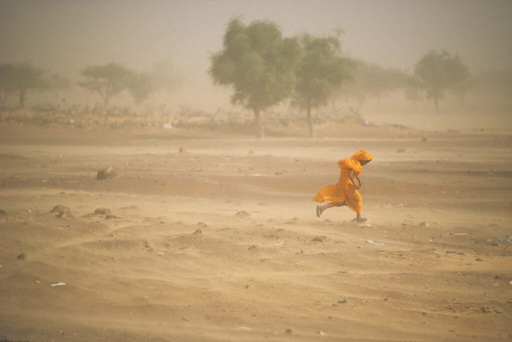 Person in orange robe walking through swirling dust storm in barren sandy landscape wallpaper
