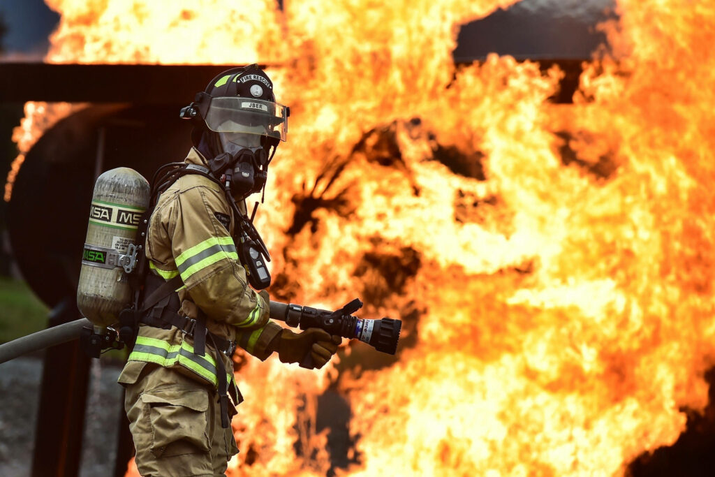 Fireman in full gear battles intense blaze with hose against towering flames wallpaper background