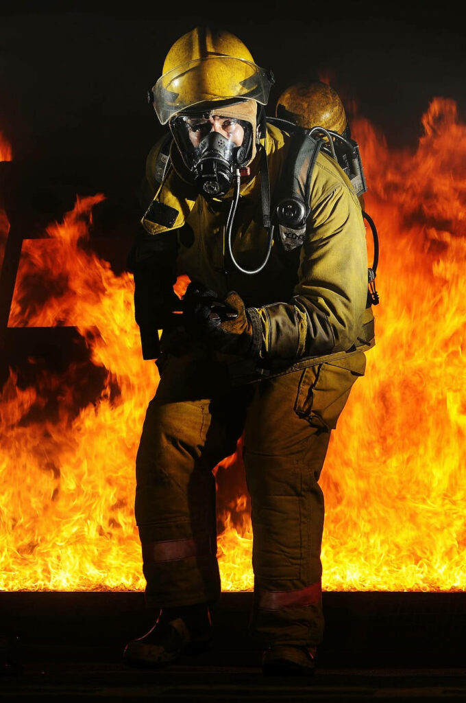 Firefighter in full gear with breathing apparatus emerging from intense flames showing courage Wallpaper
