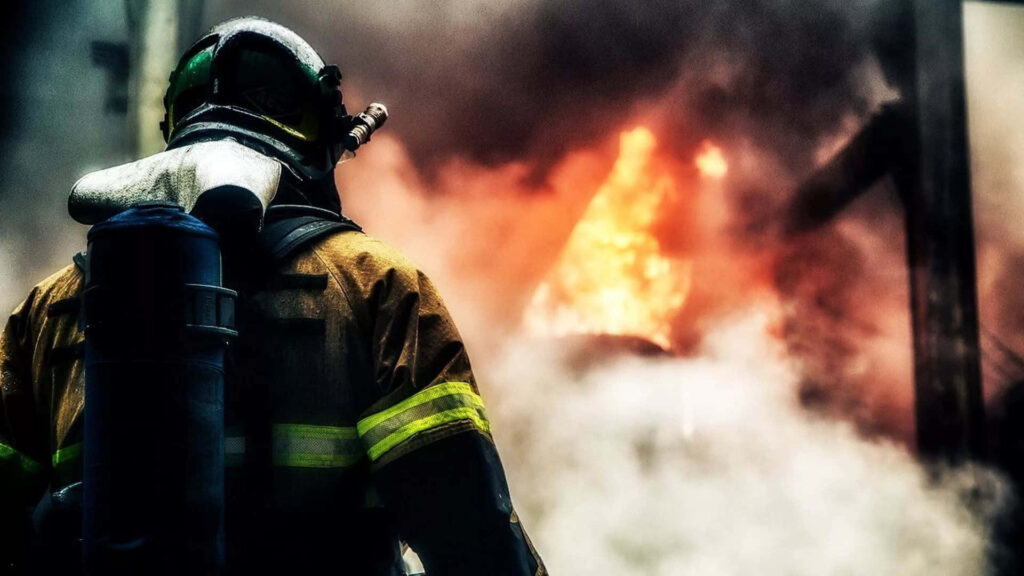 Firefighter in full gear stands amid thick smoke and flames showing courage and determination Wallpaper