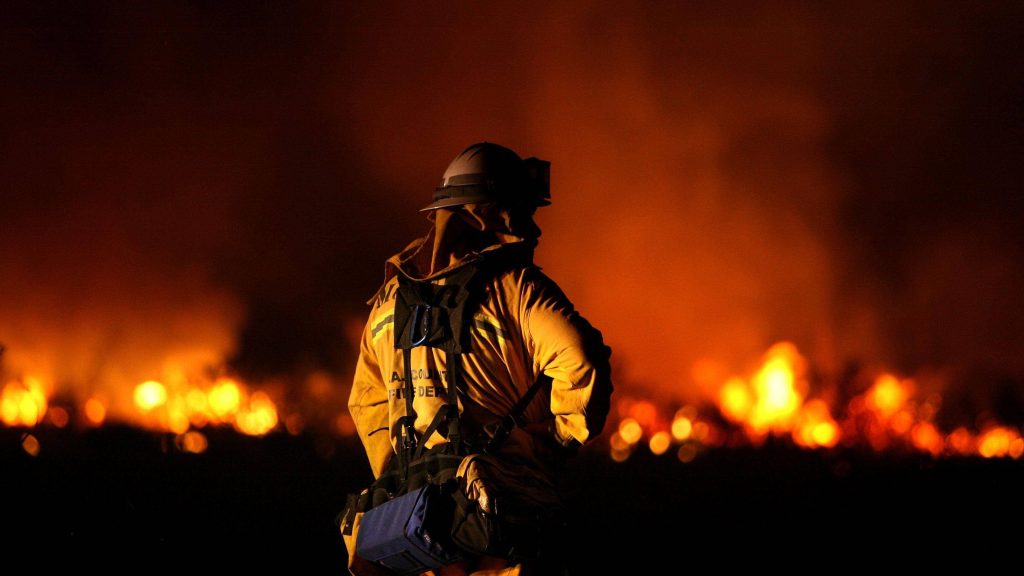 Lone firefighter in yellow gear faces massive night wildfire glowing intense flames silhouette Wallpaper