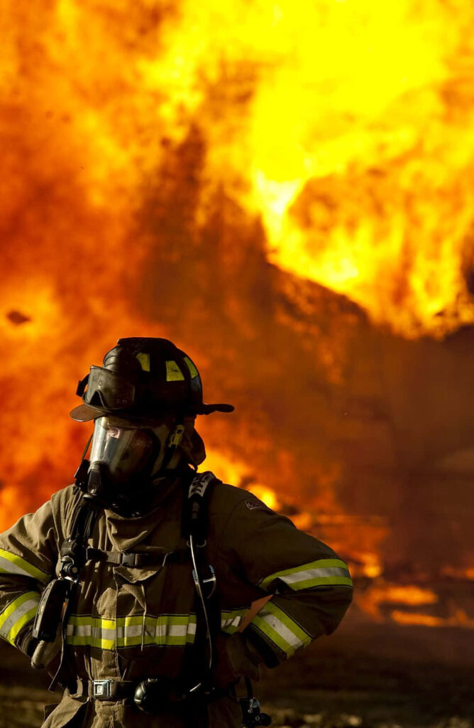 Firefighter in full gear stands boldly with blazing fire background emergency responder bravery Wallpaper