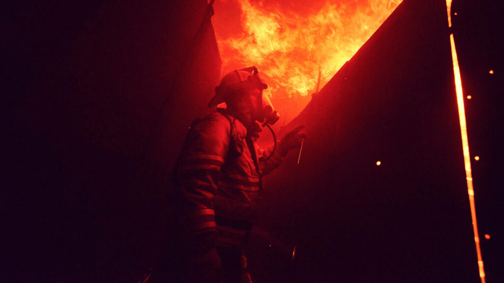 Firefighter in full gear standing boldly amid intense orange flames and smoke background wallpaper