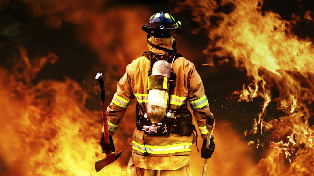 Brave firefighter in full gear walking into intense roaring flames showing courage and strength Wallpaper