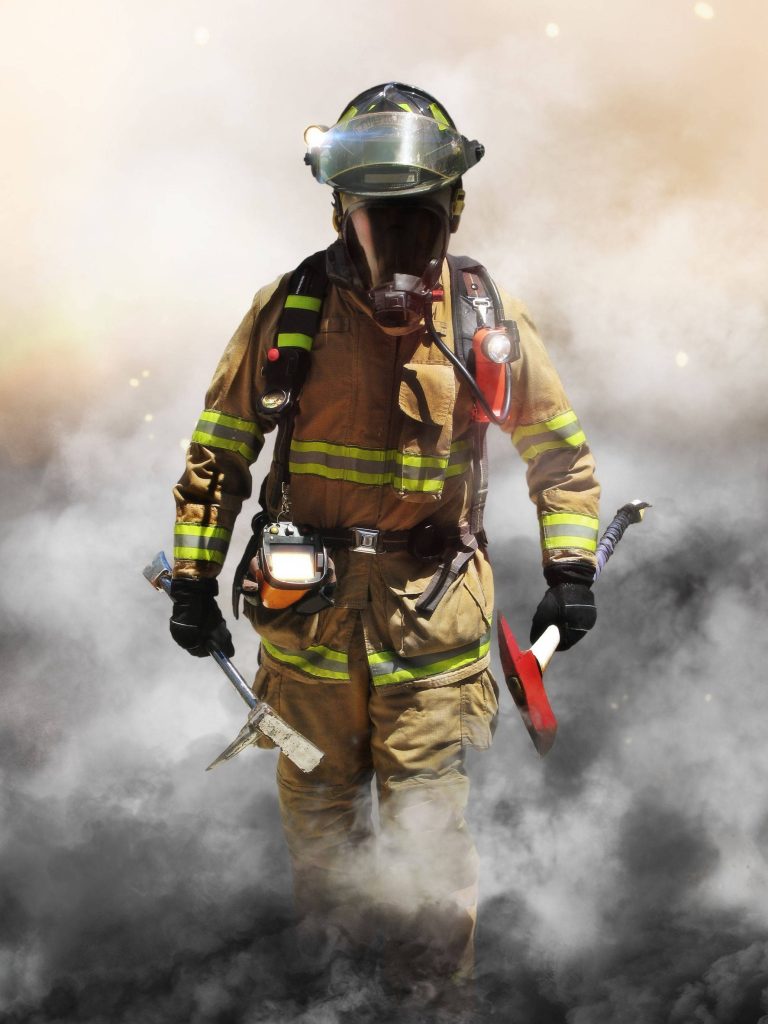 Firefighter in full gear with SCBA mask holds tools emerging from dense smoke wallpaper