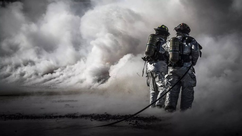 Two firefighters in MSA aluminized gear stand vigilant amidst thick smoke dramatic firefighting scene Wallpaper