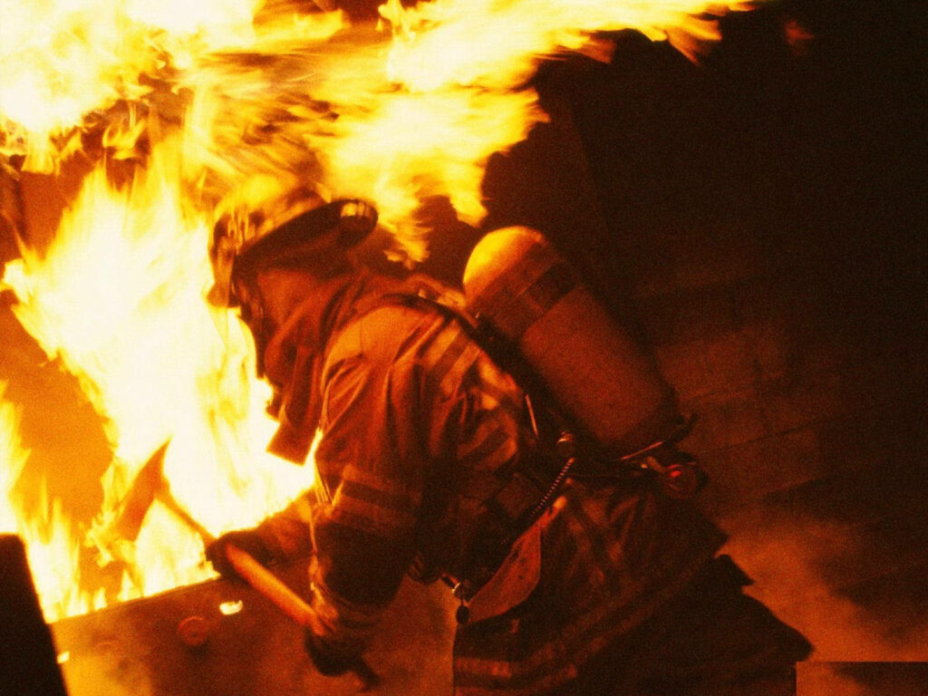 Fireman battling intense flames inside burning building surrounded by smoke and orange firelight Wallpaper