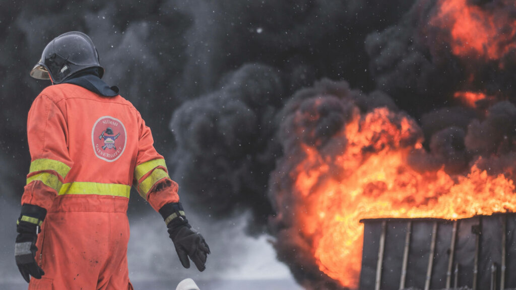 Fireman in bright orange gear stands ready amid intense flames and swirling smoke wallpaper background