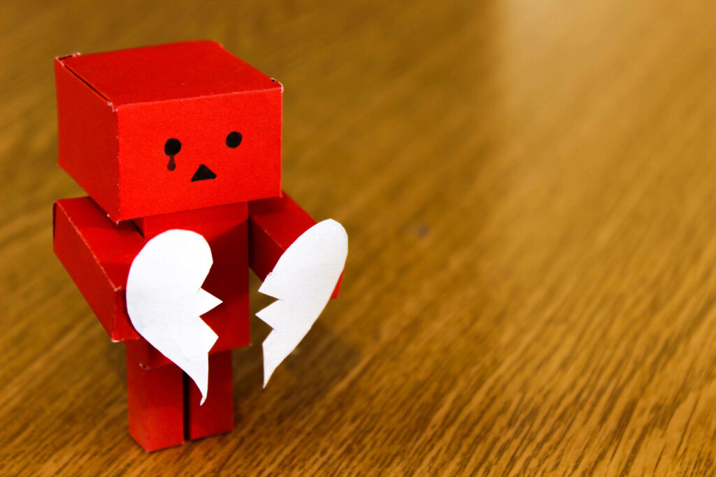 Minimalist red cardboard figure with sad expression holding broken white heart on wood background Wallpaper
