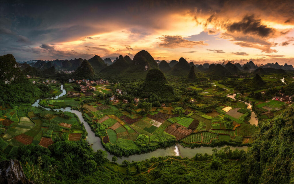 A Serene Chinese Countryside at Sunset: An Idyllic Village Amidst Vibrant Farmlands, Scenic River, and Majestic Mountain Peaks Wallpaper