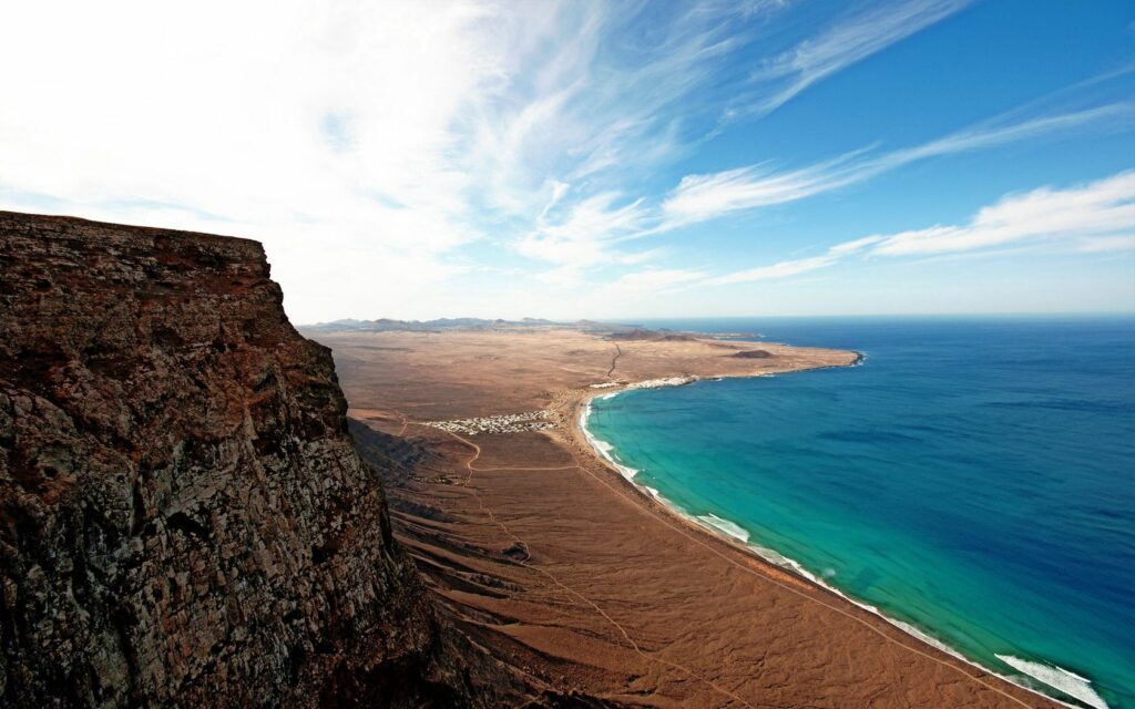 Rugged cliff with sandy shore and turquoise sea under vibrant sky coastal landscape wallpaper