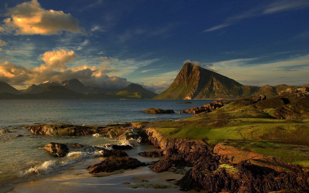 Breathtaking coastal landscape with mossy slopes and mountains under golden cloudy sky wallpaper