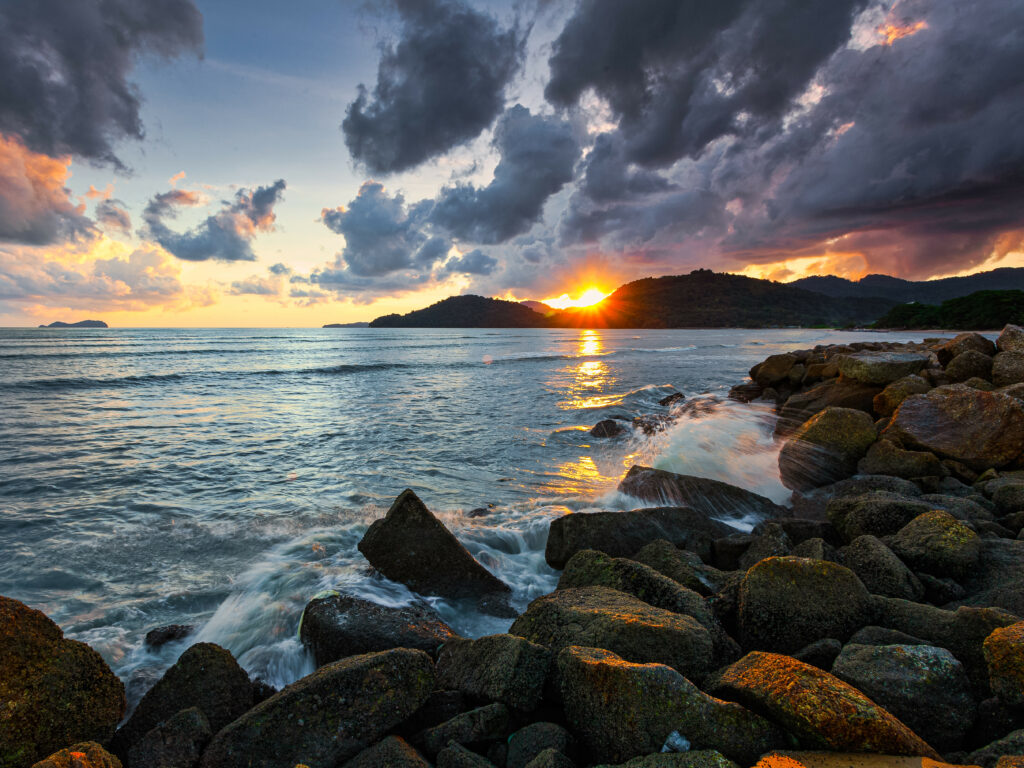 Breathtaking coastal sunset with golden rays over tranquil sea rugged stones and silhouetted hills Wallpaper