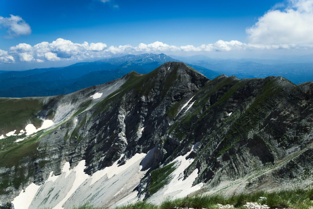 Italian mountain peak with rugged cliffs snow patches green grass and vivid blue sky clouds Wallpaper