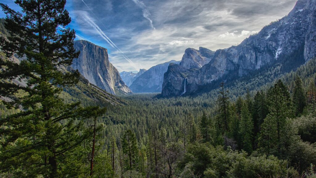 Breathtaking mountain landscape with rocky cliffs lush valley dense forest and dramatic sky sunlight Wallpaper