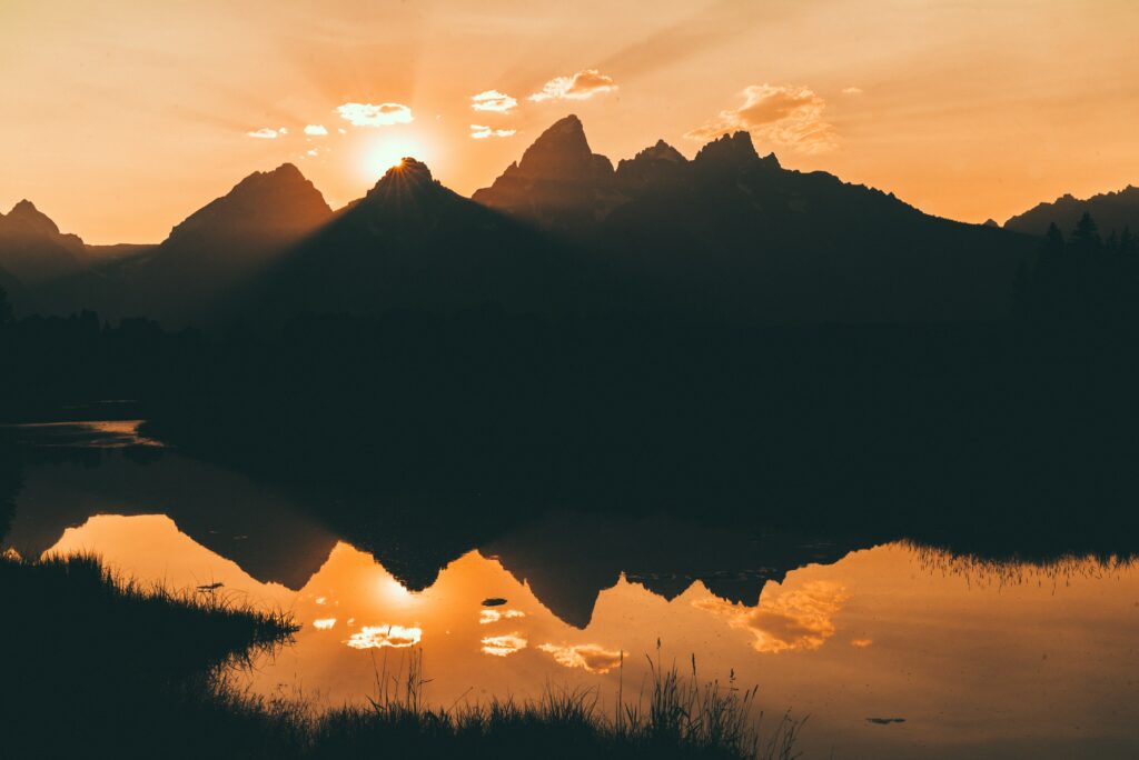 Sunset over rugged mountain peaks with glowing sky and vibrant orange reflection on calm water Wallpaper