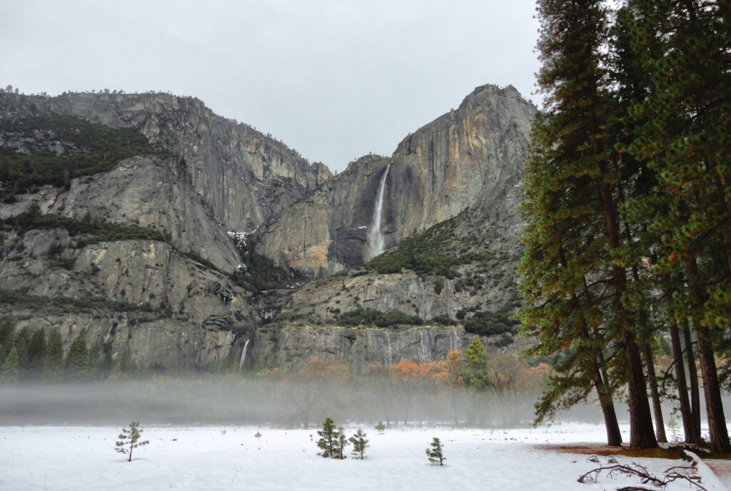 Snowy mountain cliffs with cascading waterfall evergreen trees and fog under overcast sky wallpaper