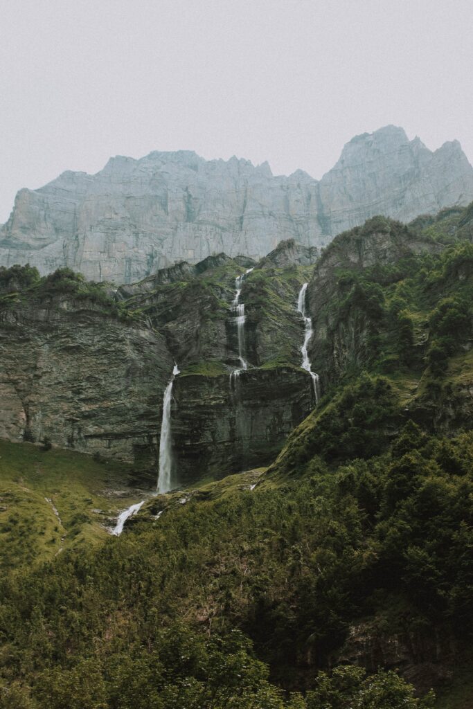 Towering mountains with cascading waterfalls over rocks surrounded by lush green forest wallpaper