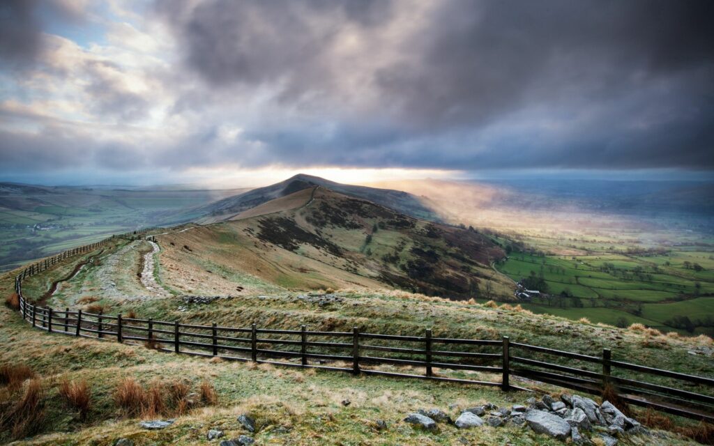 Rolling green hills under dramatic sky with sunlight and rustic wooden fence landscape wallpaper
