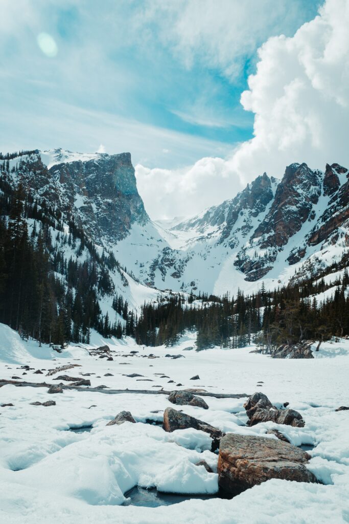 Snowy mountain landscape with rugged rocks evergreen trees and vibrant blue sky with clouds Wallpaper