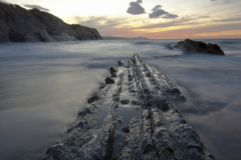 Sunset over rugged rocks with misty sea and gentle waves creating a serene ocean landscape Wallpaper