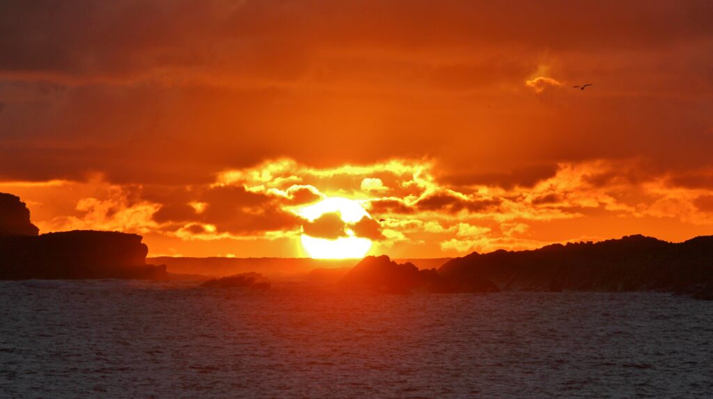 Sunset over rugged sea rocks with orange and gold sky reflecting on calm ocean at dusk wallpaper